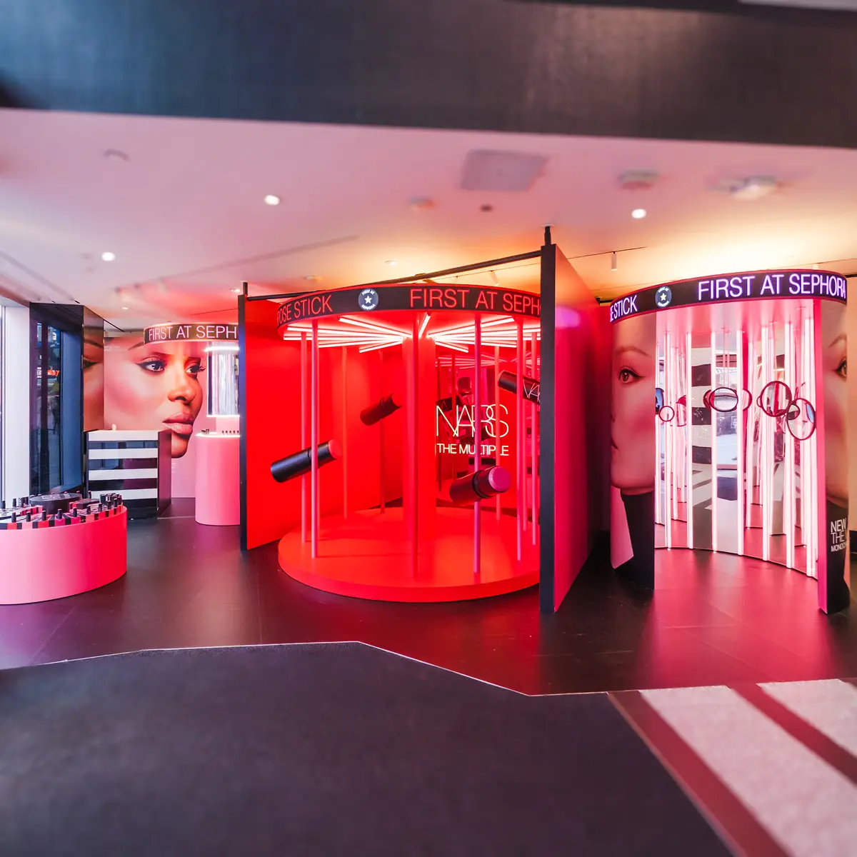 A brightly lit Sephora store display, designed and produced by Arsenal, features red and pink circular booths with bold makeup images and NARS branding, highlighting lipsticks and beauty products for a captivating brand experience under the sign “FIRST AT SEPHORA.”.