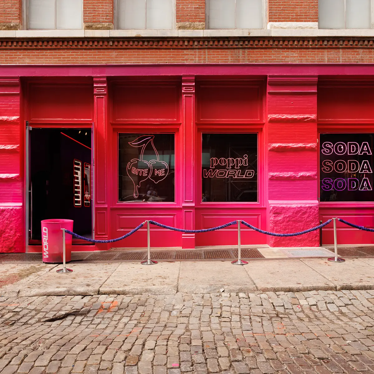 A bright pink storefront—design and produced by Arsenal—with large windows displaying neon signs that read BITE ME, poppi WORLD, and SODA SODA SODA. A rope barrier lines the entrance, promising a unique brand experience on the cobblestone sidewalk.