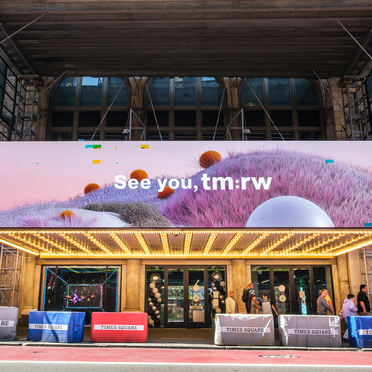 A large digital billboard in Times Square reads See you, tm:rw above an entrance with bright marquee lights. The billboard, design and produced by Arsenal, displays a futuristic landscape with abstract purple and orange shapes. Pedestrians walk below.